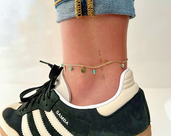 May include: Gold ankle chain with turquoise beads and small gold discs. The chain is worn on a person's ankle with a black and white sneaker. The numbers 1995 are written on the ankle.