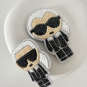 May include: Two handcrafted beaded brooches depicting a stylized figure with white hair, black sunglasses, and a black suit. The designs are created using small white, beige, and black beads, with silver-toned thread outlining the edges of each piece.