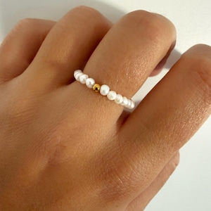 May include: A ring featuring a continuous band of small, white, irregular freshwater pearls interspersed with a single, smooth, polished gold-tone spherical bead centered among the pearls, creating a delicate and simple circular jewelry piece.