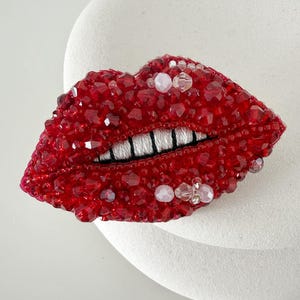 May include: A brooch shaped like a pair of lips, densely covered in faceted red beads and crystals. The design features a row of white thread teeth with black vertical stitching to define the individual teeth, creating a textured, sparkling appearance.