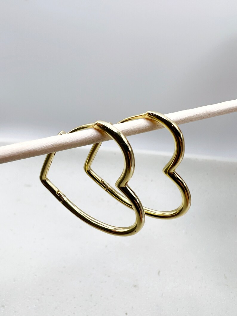 Peut inclure: Une paire de boucles d'oreilles cr&eacute;oles en forme de c&oelig;ur dor&eacute;es. Les boucles d'oreilles sont suspendues &agrave; un b&acirc;tonnet blanc.