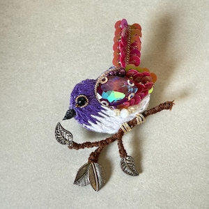 May include: A handcrafted bird brooch with a purple and white embroidered body, featuring a large iridescent gemstone and a tail of pink sequins. The bird is perched on a brown twig with metal leaf accents, a whimsical accessory.
