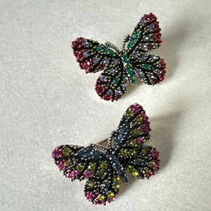 Może przedstawiać: Dwa pierścionki w kształcie motyli. Każdy pierścionek ma szczegółowy wzór z kolorowymi kamieniami szlachetnymi. Skrzydła zdobione są czerwonymi, zielonymi, fioletowymi, niebieskimi i żółtymi kamieniami. Pierścionki są na jasnoszarym tle.