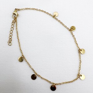 May include: Gold-colored anklet featuring a delicate chain design. The anklet is adorned with small, round, flat disc charms evenly spaced along the chain. The adjustable clasp and chain are visible, set against a white background.