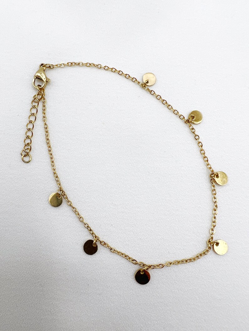 May include: Gold-colored anklet featuring a delicate chain design. The anklet is adorned with small, round, flat disc charms evenly spaced along the chain. The adjustable clasp and chain are visible, set against a white background.