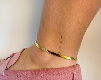 May include: A gold-toned herringbone chain anklet worn around the ankle. The anklet features a flat, flexible, and reflective metal link design that sits flush against the skin. The jewelry is positioned above a pink suede loafer shoe.