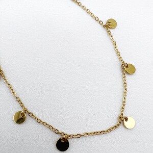 May include: A delicate gold-colored necklace featuring a fine chain adorned with small, flat, circular charms. The charms are evenly spaced along the chain, creating a minimalist and elegant design. The necklace is displayed against a white background.