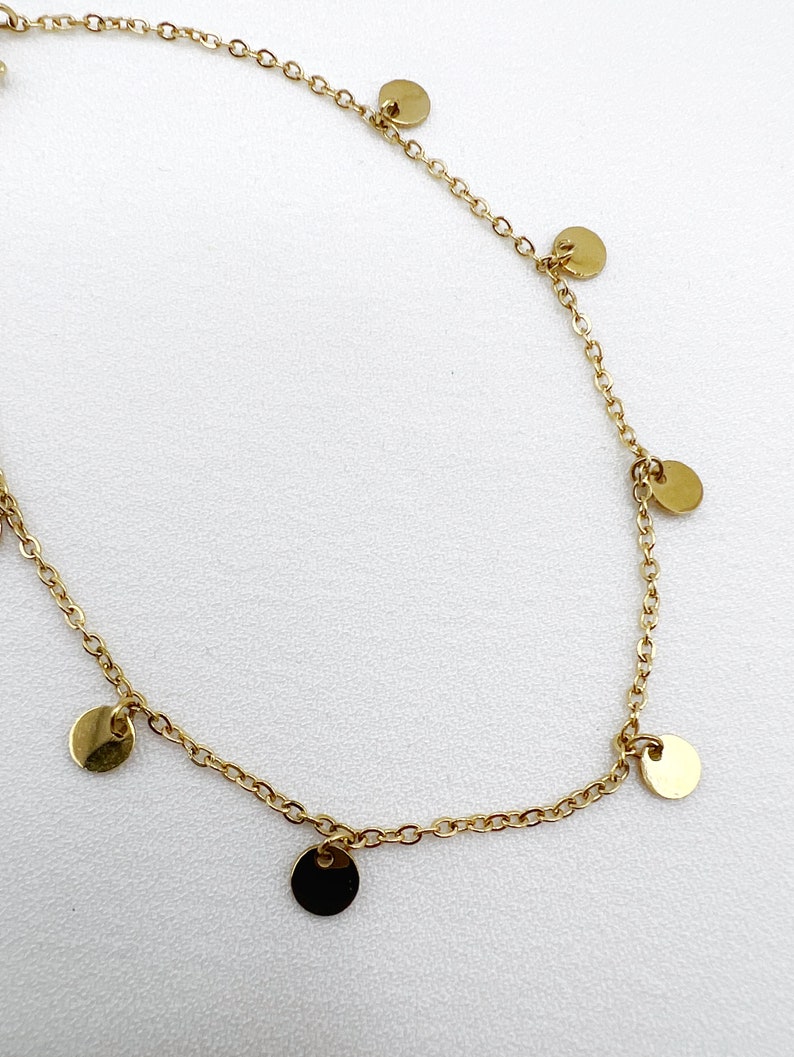 May include: A delicate gold-colored necklace featuring a fine chain adorned with small, flat, circular charms. The charms are evenly spaced along the chain, creating a minimalist and elegant design. The necklace is displayed against a white background.