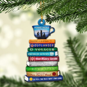 May include: A Christmas ornament shaped like a stack of books with the titles "Dragonfly in Amber", "Outlander", "Voyager", "Drums of Autumn", "The Fiery Cross", "A Breath of Snow and Ashes", "An Echo in the Bone", "Written in My Own Heart's Blood", and "Go Tell the Bees That I Am Gone". The ornament also features a teacup with a silhouette of two people standing on a hill.