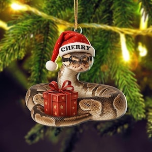 Pode incluir: Um enfeite de Natal com uma cobra usando um gorro de Papai Noel com o nome "Cherry". A cobra está enrolada em uma caixa de presente vermelha com estrelas douradas. O enfeite está pendurado em frente a uma árvore de Natal desfocada.