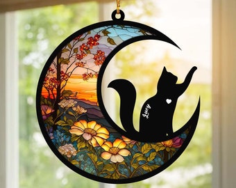 Può includere: Un ornamento a forma di luna in vetro colorato con una silhouette di gatto nero e il nome "Lucy" inciso. La luna è circondata da fiori colorati e una scena di tramonto.