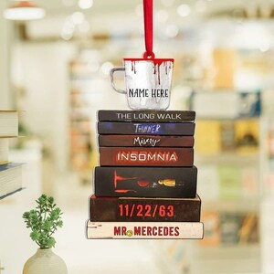 May include: A stack of books with the titles "The Long Walk", "Thinner", "Misery", "Insomnia", "11/22/63", and "Mr. Mercedes". A white coffee mug with red dripping paint and the text "NAME HERE" is on top of the stack of books.