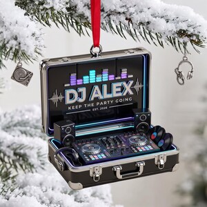 Puede incluir: Un adorno en forma de maletín negro que contiene un controlador de DJ, altavoces y auriculares. La tapa interior muestra un ecualizador gráfico, el texto "DJ ALEX KEEP THE PARTY GOING EST. 2026" y una cinta roja para colgarlo.