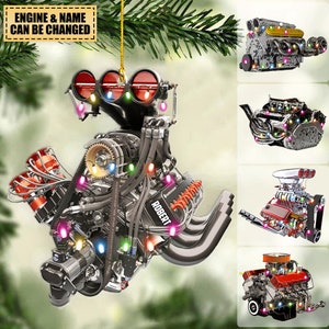 Puede incluir: Un adorno navideño con forma de motor de coche con luces de colores. El motor está hecho de metal y tiene un acabado negro y plateado. El adorno está colgado de una rama de árbol. El texto "ROBERT" es visible en el motor.