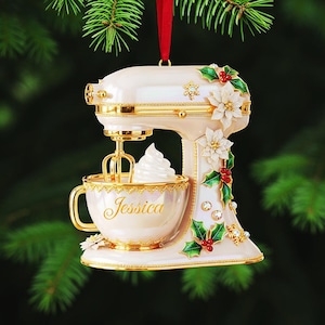 May include: A cream-colored mixer Christmas ornament with gold accents, a red ribbon, and a cup with the name "Jessica". The ornament is decorated with white flowers, green holly leaves, and red berries. The mixer has a whisk and a dollop of whipped cream.