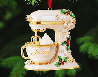 May include: A cream-colored mixer Christmas ornament with gold accents, a red ribbon, and a cup with the name "Jessica". The ornament is decorated with white flowers, green holly leaves, and red berries. The mixer has a whisk and a dollop of whipped cream.
