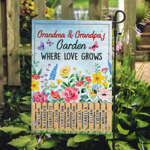 May include: A colorful garden flag with the text "Grandma & Grandpa's Garden Where Love Grows". The flag features a picket fence with names of children written on each picket. The flag is decorated with butterflies and flowers.