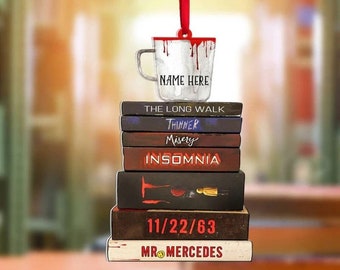 May include: A white coffee mug with red dripping paint and the text "NAME HERE" hanging from a red ribbon. The mug is stacked on top of a stack of books with the titles "The Long Walk", "Thinner", "Misery", "Insomnia", "11/22/63", and "Mr. Mercedes".