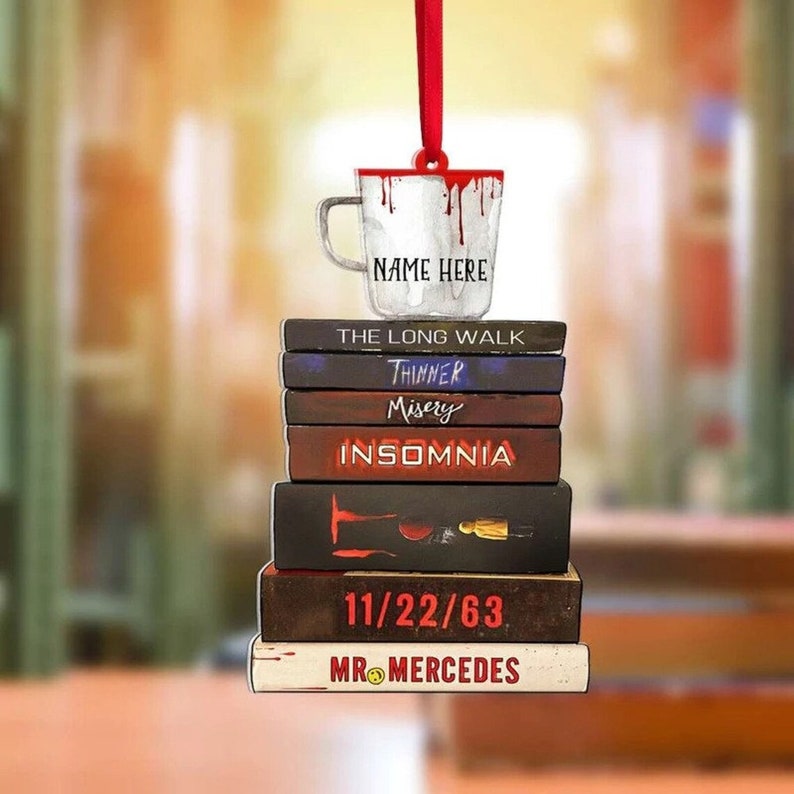 May include: A white coffee mug with red dripping paint and the text "NAME HERE" hanging from a red ribbon. The mug is stacked on top of a stack of books with the titles "The Long Walk", "Thinner", "Misery", "Insomnia", "11/22/63", and "Mr. Mercedes".