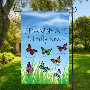 May include: A colorful garden flag with a blue sky background and white clouds. The text "Grandma's Butterfly Kisses" is printed in black. There are six butterflies with different colors and patterns, each with a name printed below: Keeleigh, Nale, Emmaleigh, Mason, Addison, Daniel, and Jacob.