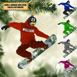 May include: A red, white, and black snowboard ornament with a snowboarder wearing a red hoodie, black helmet, and goggles. The ornament is personalized with the name "WILLIAM" and the number "23".