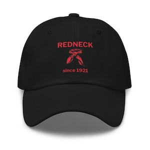 May include: Black baseball cap with the word "REDNECK" in red block letters above a red graphic of a neckerchief. The text "since 1921" is below the graphic. The cap has a curved brim and a fabric strap closure.