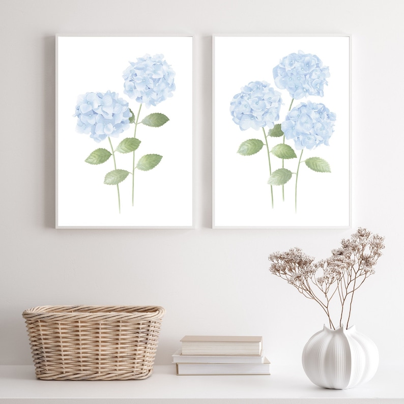 May include: Two framed prints of watercolor paintings of blue hydrangeas. The prints are hung on a white wall above a wicker basket and a white vase with dried flowers.