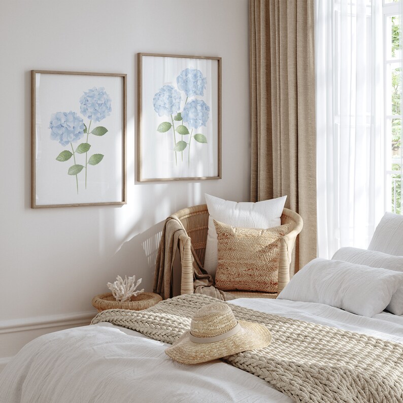May include: A white bed with a beige knitted throw blanket and a straw hat. A wicker chair with a white pillow and a beige pillow is in the background. Two framed watercolor paintings of blue hydrangeas are on the wall.