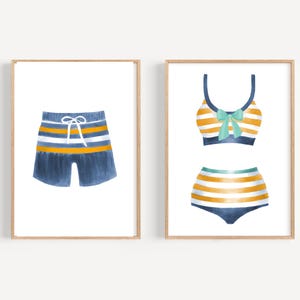 May include: Two framed watercolor prints. One features blue swim trunks with white and yellow stripes and a drawstring. The other shows a two-piece swimsuit with yellow and white stripes, a blue bow, and blue trim.
