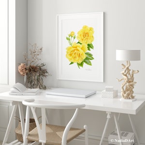 May include: A watercolor painting of two yellow roses with green leaves in a white frame. The painting is hanging on a white wall above a white desk with a laptop, a book, and a lamp.