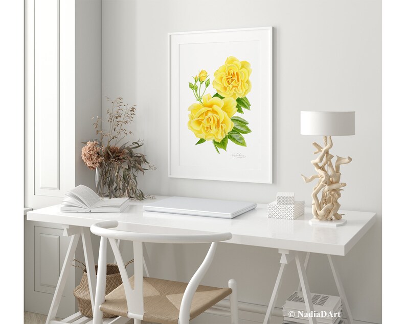 May include: A watercolor painting of two yellow roses with green leaves in a white frame. The painting is hanging on a white wall above a white desk with a laptop, a book, and a lamp.