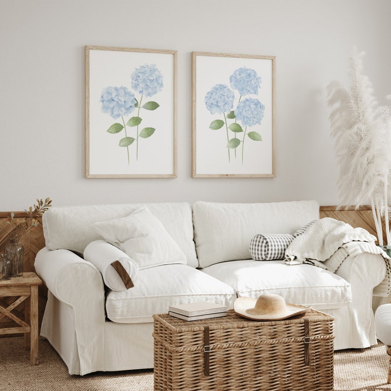 May include: Two framed watercolor prints of blue hydrangeas with green stems and leaves. The prints are hanging on a white wall above a white couch with a brown wicker storage ottoman in front of it.