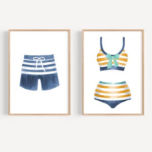 May include: Watercolor art prints of swimwear. One print features blue and white striped swim trunks with a white drawstring. The other print shows a two-piece swimsuit with yellow and white stripes, a blue bow, and blue accents.