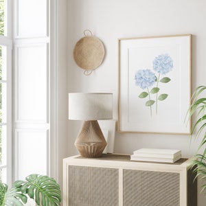 May include: A white lamp with a woven shade sits on a wooden cabinet with a woven front. A framed print of blue hydrangeas hangs on the wall above the cabinet. A woven basket hangs on the wall to the left of the print.