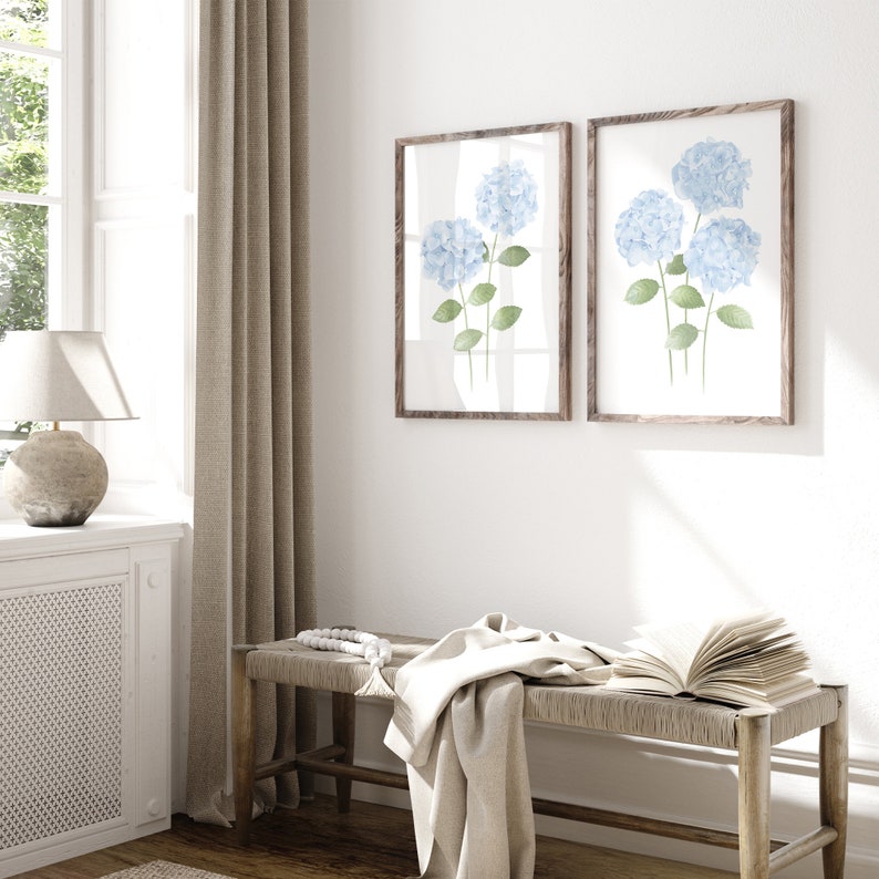 May include: Two framed watercolor prints of blue hydrangeas with green stems and leaves. The prints are hung on a white wall above a wooden bench with a white throw blanket and a book.