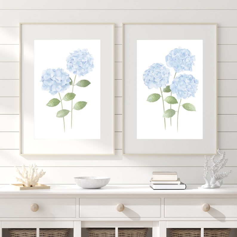 May include: Two framed watercolor prints of blue hydrangeas with green leaves on a white wood background. The prints are hanging on a white wall above a white dresser with two drawers.