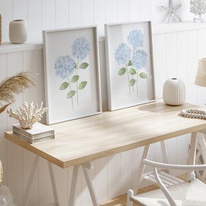 May include: Two white framed prints of watercolor blue hydrangeas on a white wood background. The prints are on a light wood desk with a white chair and a white vase with a white and brown patterned design.