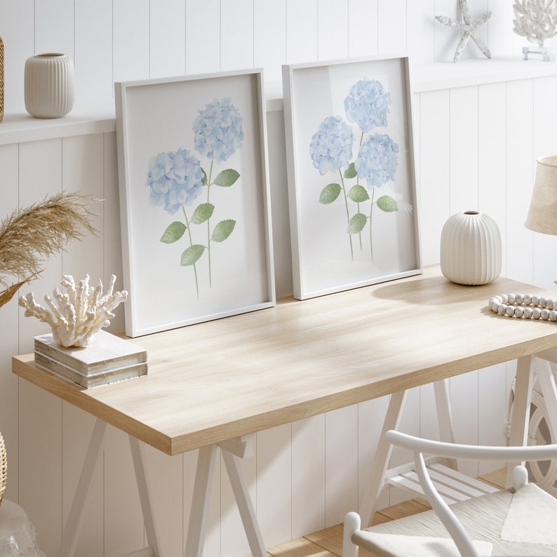 May include: Two white framed prints of watercolor blue hydrangeas on a white wood background. The prints are on a light wood desk with a white chair and a white vase with a white and brown patterned design.