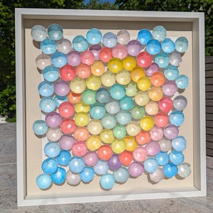 May include: A white frame with a rainbow of pastel colored eggs arranged in a circular pattern. The eggs are arranged in a gradient from light blue to pink.