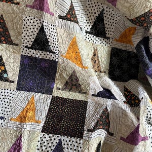 May include: A quilted Halloween-themed blanket featuring repeating square blocks. Each block displays a witch hat in various colors, including orange, purple, and black, set against patterned backgrounds of polka dots, stripes, and spiderwebs. The quilt has a cream-colored background.