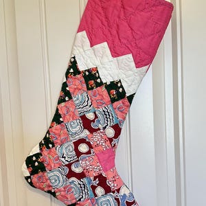May include: A pink and white patchwork Christmas stocking with a chevron pattern at the top. The stocking is made of various fabrics with floral and geometric designs.