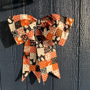 May include: A decorative fabric bow featuring a patchwork pattern of orange, black, and white squares. The design includes motifs of ghosts, pumpkins, spider webs, and checkered patterns. The bow has two long, pointed tails hanging downward.