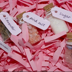 Pu&ograve; includere: Cinque piccoli flaconi di vetro con tappi di sughero, ognuno riempito con ingredienti diversi ed etichettato con etichette scritte a mano. Le etichette riportano "Lavender", "Cinnamon", "Pink Salt", "Sugar" e "Crystals". I flaconi sono su un letto di carta crespa rosa.