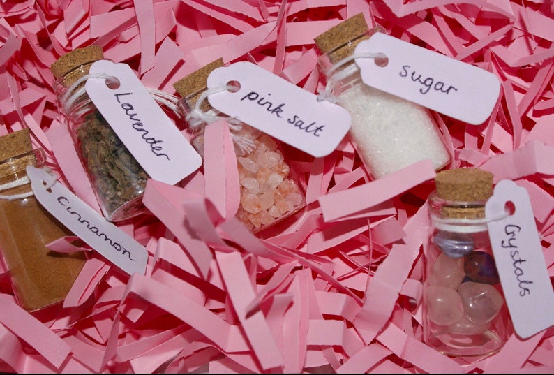Pu&ograve; includere: Cinque piccoli flaconi di vetro con tappi di sughero, ognuno riempito con ingredienti diversi ed etichettato con etichette scritte a mano. Le etichette riportano "Lavender", "Cinnamon", "Pink Salt", "Sugar" e "Crystals". I flaconi sono su un letto di carta crespa rosa.