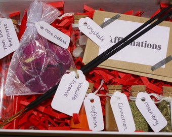 Può includere: Una scatola bianca piena di carta rossa triturata e piccole bottiglie di vetro con tappi di sughero. Le bottiglie contengono varie erbe e spezie, tra cui zafferano, cannella, rosmarino e basilico. Ci sono anche petali di rosa in un sacchetto di organza bianco, cristalli in un piccolo barattolo di vetro e due bastoncini di incenso neri con un'etichetta che dice "affermazioni".