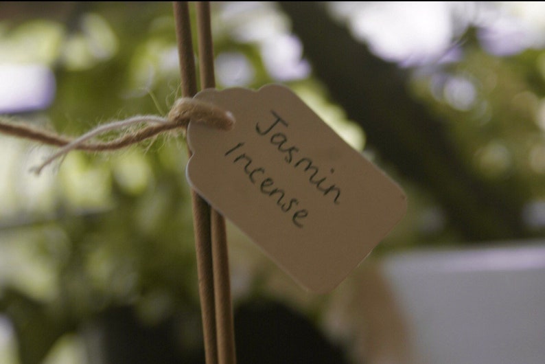 Pu&ograve; includere: Primo piano di un'etichetta di carta marrone legata con spago a due bastoncini di incenso marroni. L'etichetta recita "Jasmin Incense".