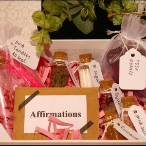 Pu&ograve; includere: Una scatola piena di vari oggetti per un rituale o un incantesimo, tra cui candele rosa, petali di rosa, zucchero, cannella, sale rosa e una piccola bottiglia di cristalli. La scatola contiene anche una carta con la parola "Affirmations" scritta su di essa.