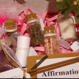 Pu&ograve; includere: Una scatola regalo rosa e bianca piena di piccoli barattoli di vetro contenenti cristalli, sale e altri oggetti. La scatola include anche un pezzo di carta arrotolato e un bastoncino di legno marrone. La scatola ha un'etichetta che dice "Affirmations".