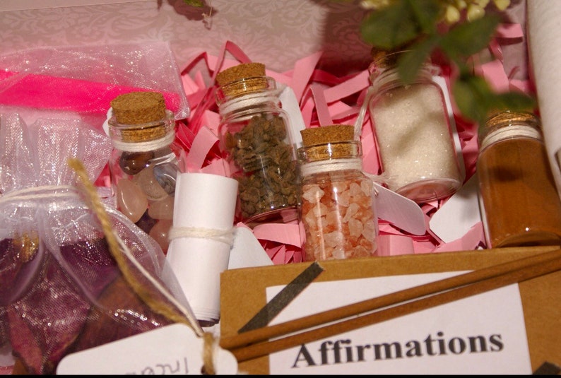 Pu&ograve; includere: Una scatola regalo rosa e bianca piena di piccoli barattoli di vetro contenenti cristalli, sale e altri oggetti. La scatola include anche un pezzo di carta arrotolato e un bastoncino di legno marrone. La scatola ha un'etichetta che dice "Affirmations".