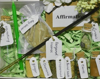 Può includere: Una scatola bianca piena di carta verde triturata contiene una varietà di oggetti per un rituale o un incantesimo. Gli oggetti includono una candela verde, una piccola borsa di foglie di alloro, una piccola bottiglia con una chiave, una piccola bottiglia di cristalli, un bastoncino di incenso al bergamotto e sei piccole bottiglie di erbe etichettate come "zenzero", "camomilla", "menta", "semi di finocchio", "chiodi di garofano" e "cannella". È inclusa anche una carta con la parola "Affermazione".